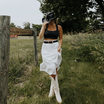 Woman in a black top, white skirt, and white boots standing in a grassy field with a wooden fence and trees in the background.