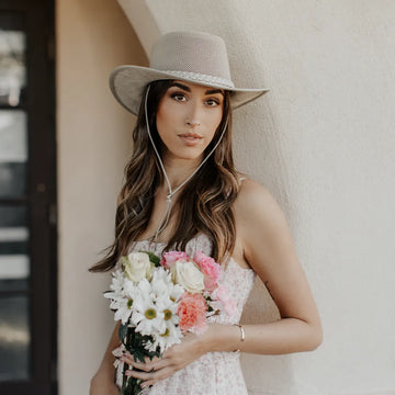 Woman wearing a wide-brimmed hat and holding flowers against a neutral background