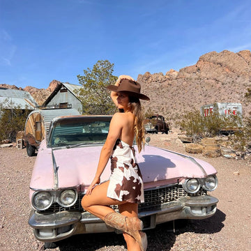 Woman in a cowboy hat and dress posing on an old pink car in a desert setting