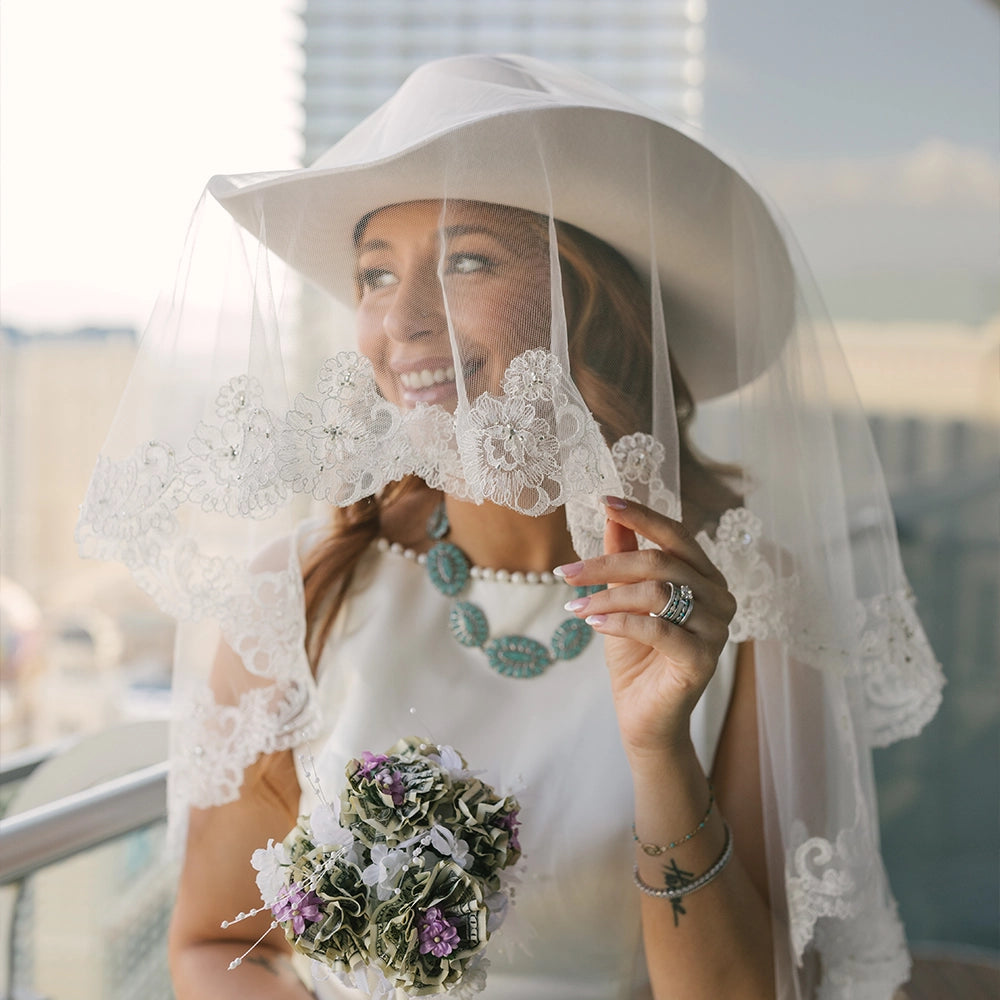 Woman in a white dress with a lace veil and bouquet, smiling outdoors.