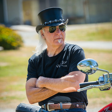 Man with long white hair wearing a black top hat and sunglasses, standing next to a motorcycle.