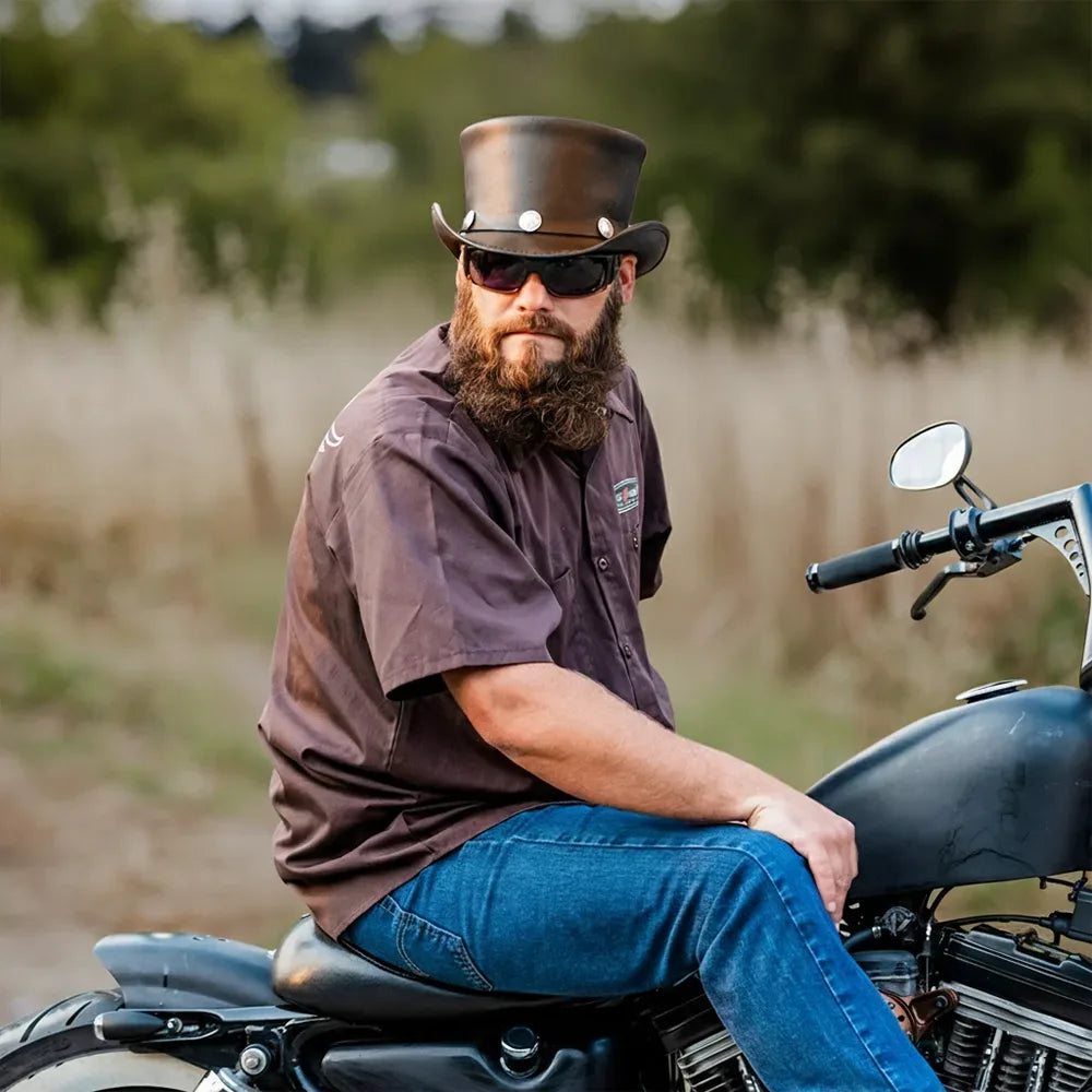 Man with a beard wearing sunglasses and a top hat sitting on a motorcycle in a field.