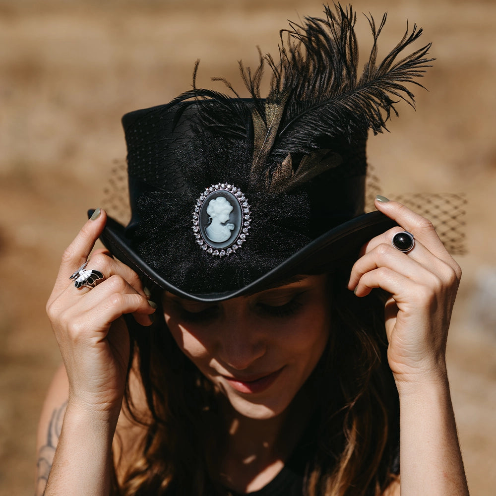 Person wearing a black hat with feather and cameo brooch, holding it with both hands.