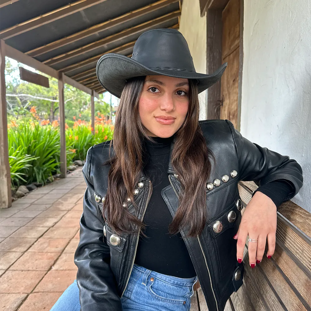 Woman wearing a black leather jacket and hat leaning against a wooden structure outdoors.