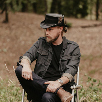 Man wearing a black cowboy hat with a feather, sitting outdoors in a natural setting.