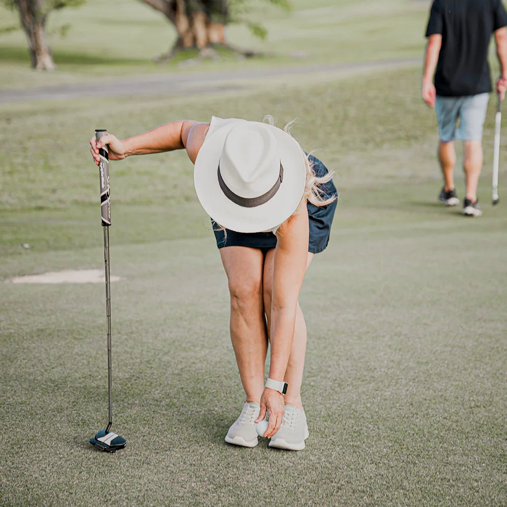 Person on a golf course wearing a hat and shorts, with another person in the background.