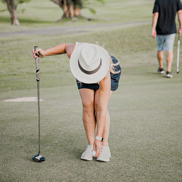 Person on a golf course wearing a hat and shorts, with another person in the background.