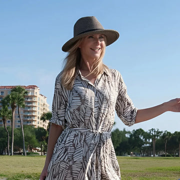 Woman in a patterned dress and hat standing in a park with palm trees and buildings in the background.