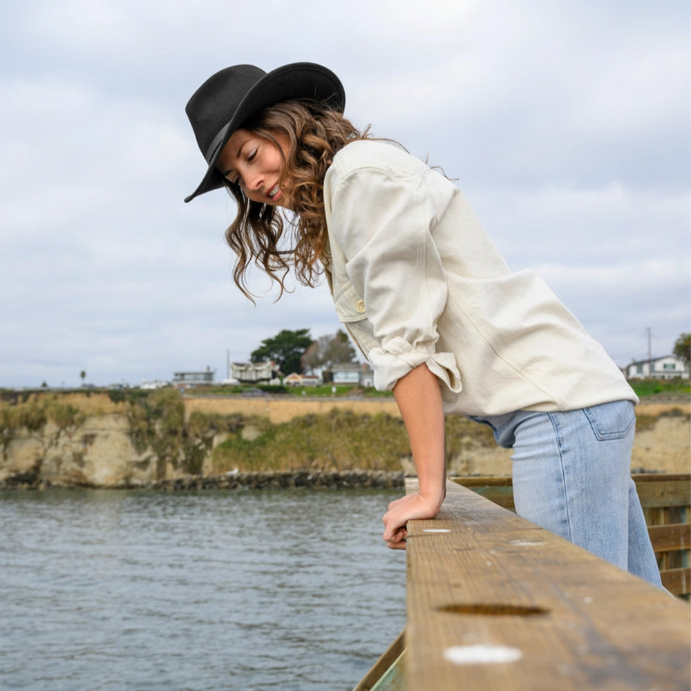 Woman wearing a black hat and white shirt leaning over a wooden railing by a body of water.