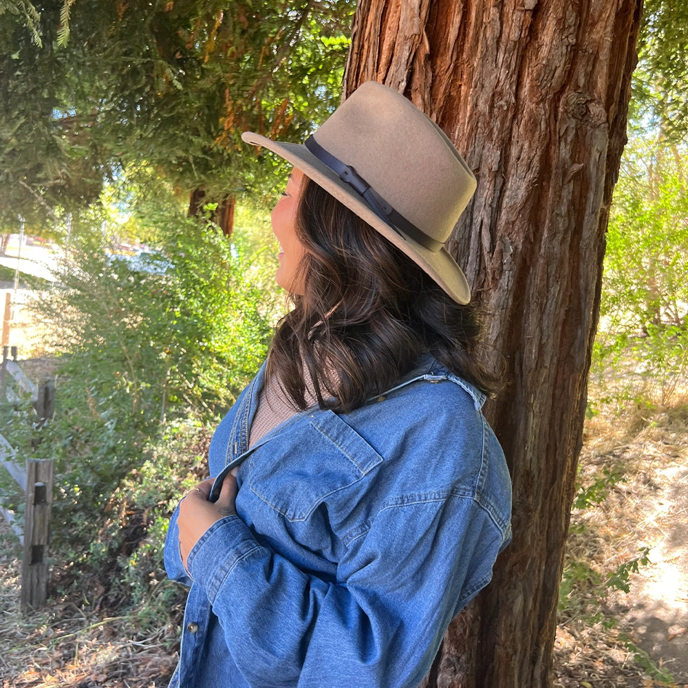 Person wearing a beige hat and blue denim jacket leaning against a tree in a forest setting