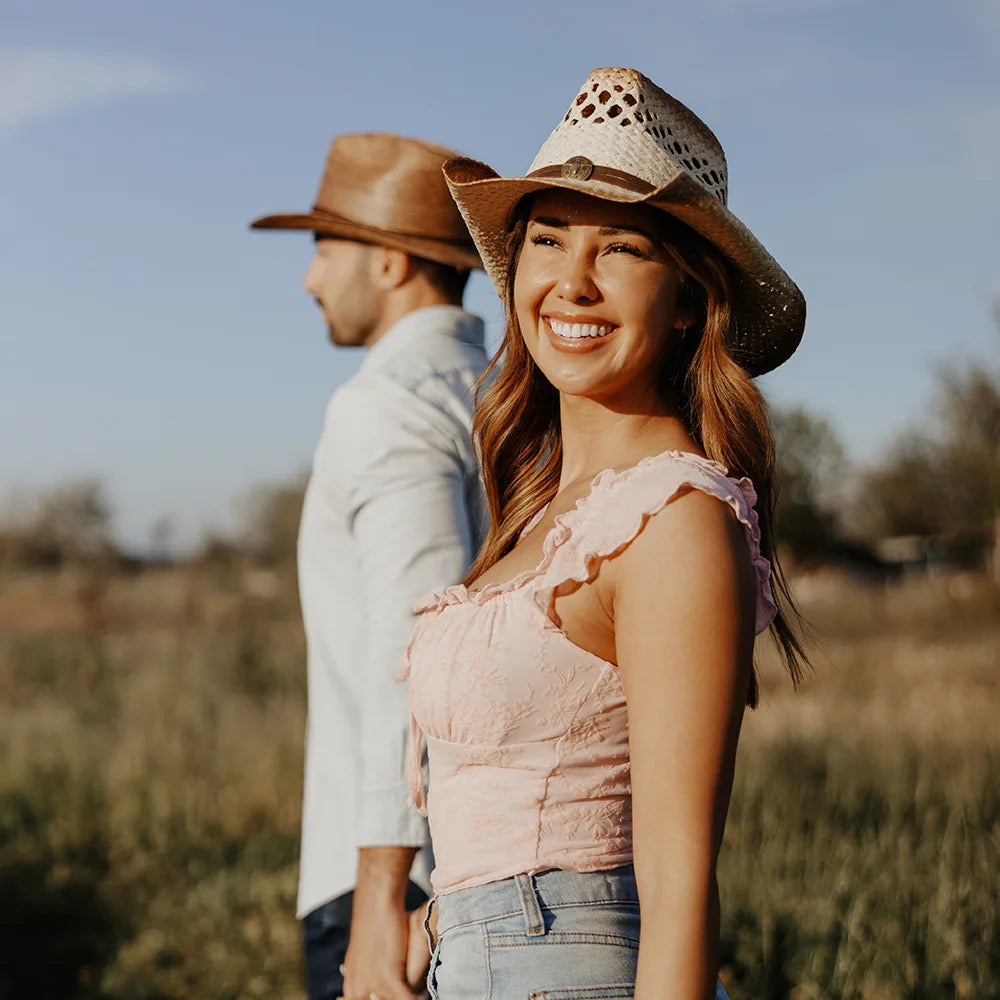 Woman wearing a cowboy hat in a field with a man partially visible behind her.