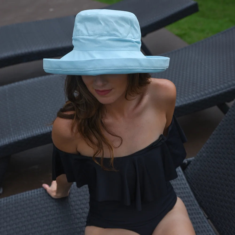 Woman wearing a light blue bucket hat and black off-shoulder swimsuit sitting on a wicker chair.