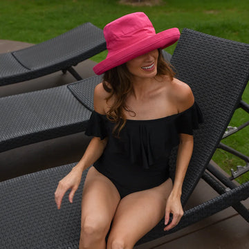 Woman in a black swimsuit and pink hat sitting on a lounge chair outdoors.