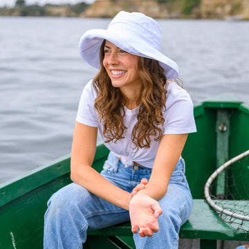 Woman sitting in a green boat by a body of water, wearing a white sun hat and white shirt.