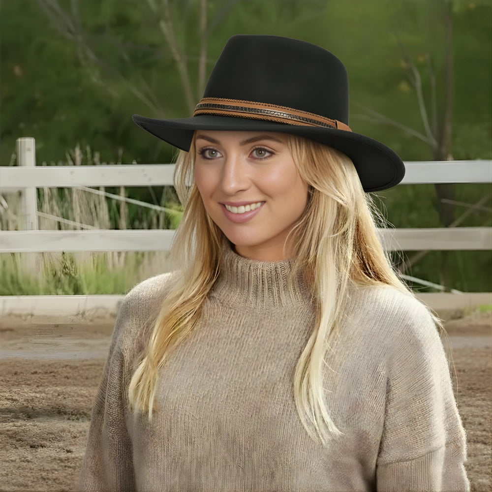 Woman wearing a black hat with a brown band, standing in an outdoor setting with a white fence and greenery.