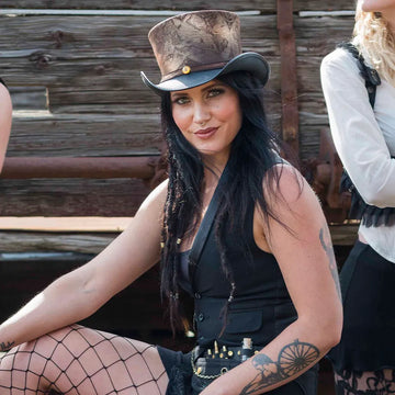 Woman wearing a top hat and black outfit with tattoos, sitting outdoors.