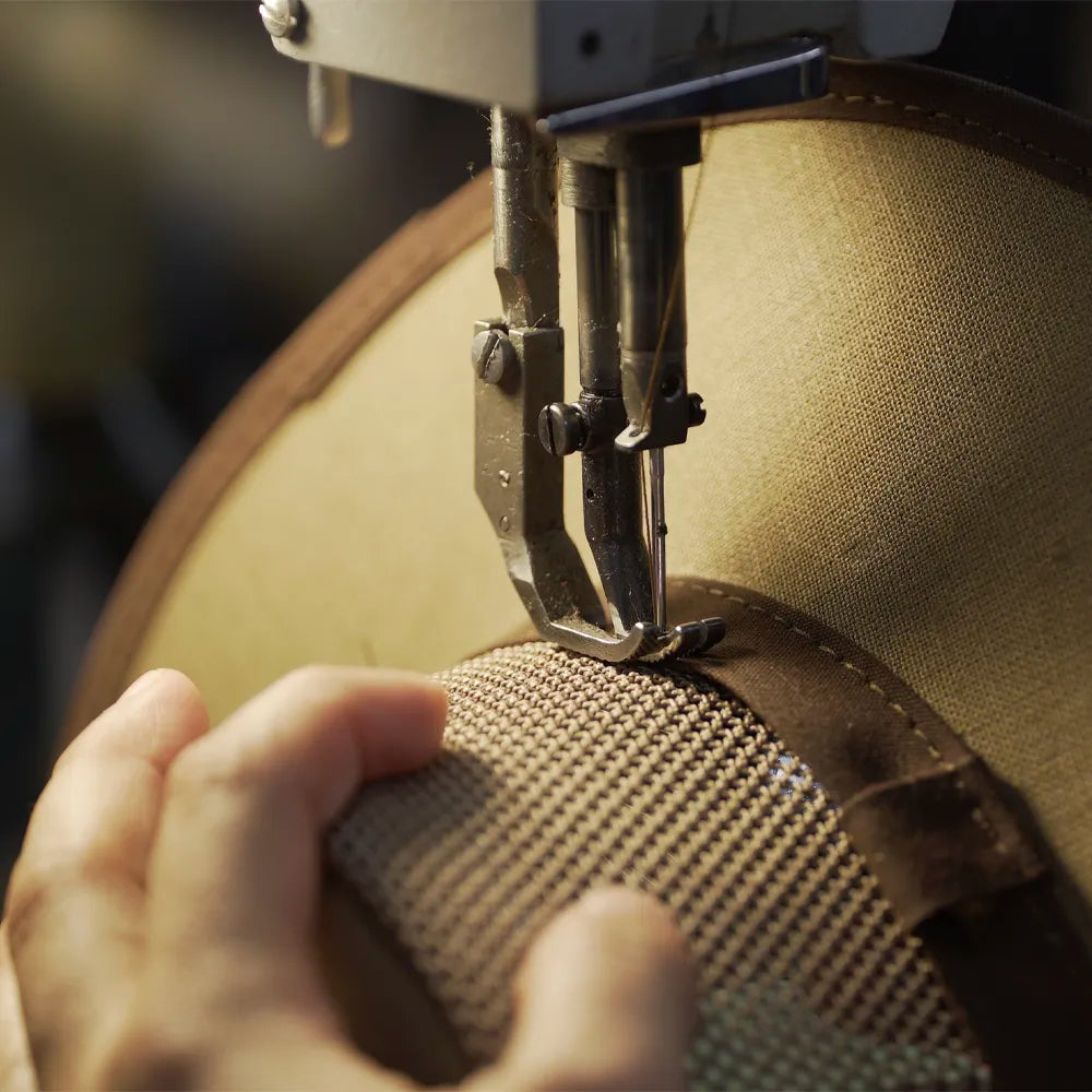 A close-up of a sewing machine stitching a beige woven hat with a brown leather trim, with a person's hand guiding the fabric.
