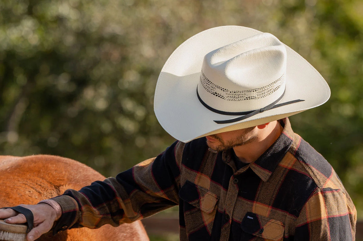 man brushing a horse wearing a plaid flannel and wearing a white straw 