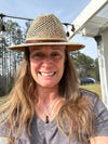 A smiling woman outdoors wearing a woven straw fedora hat with a brown band and a gray V-neck t-shirt.