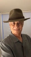 A man wearing a wide-brimmed olive green fedora hat and a matching gray-green collared shirt, photographed indoors against a light-colored wall.
