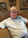 A man wearing a light beige straw hat and a light blue short-sleeve button-up shirt, sitting on a brown couch in a living room.