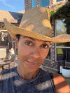 A woman wearing a natural straw cowboy hat with a wide brim, photographed outdoors in front of a brick house.