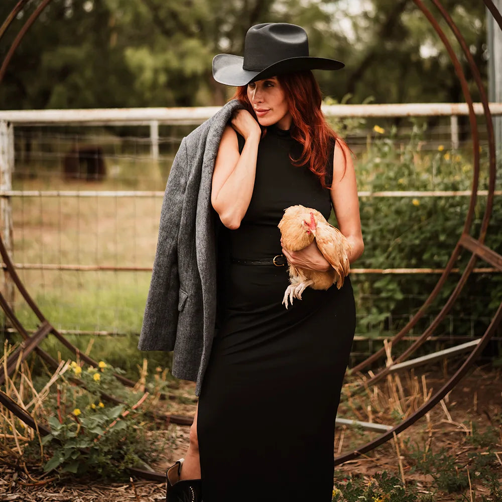 Woman in black dress and hat holding a chicken outdoors with a rustic background