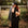 Woman in black dress and hat holding a chicken outdoors with a rustic background