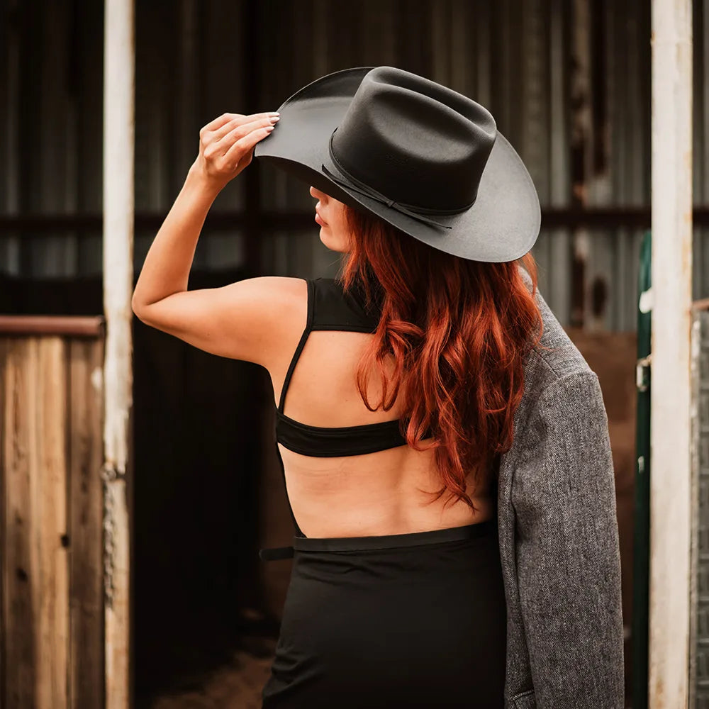 Woman wearing a black hat and black top with a gray coat, standing in front of a wooden door.