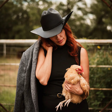 Woman wearing a black hat and gray coat holding a chicken outdoors.