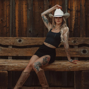 Woman in black outfit and cowboy hat sitting on a wooden bench with a rustic background