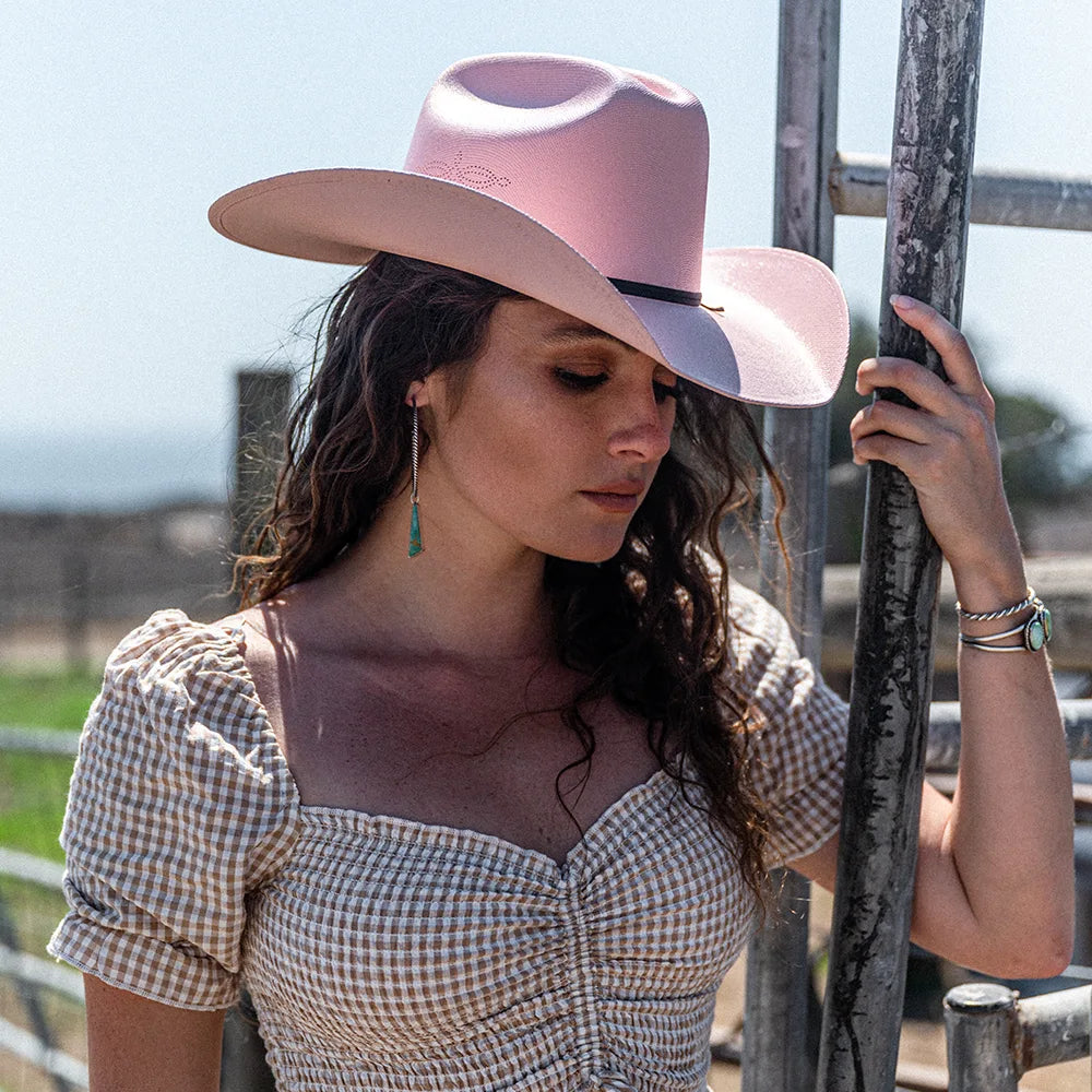 Woman wearing a pink cowboy hat and checkered top, standing near a wooden fence.