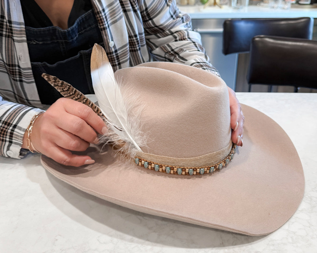 Hands adjusting feather accents on a beige felt wide-brim western hat.