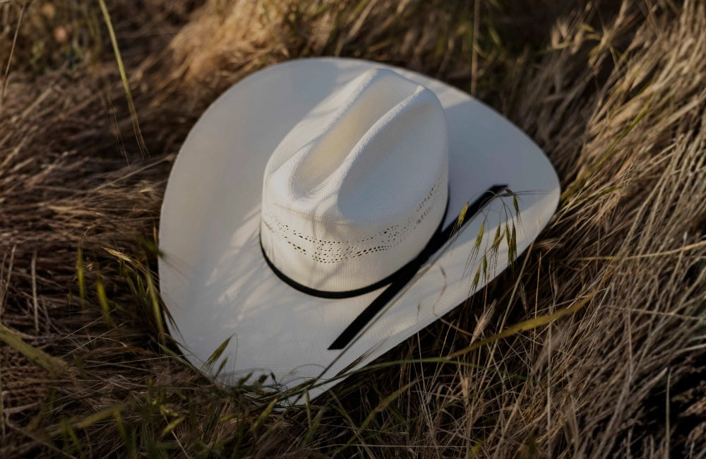 White woven straw cowboy hat with a black hatband and ventilated crown, resting in tall dry grass.