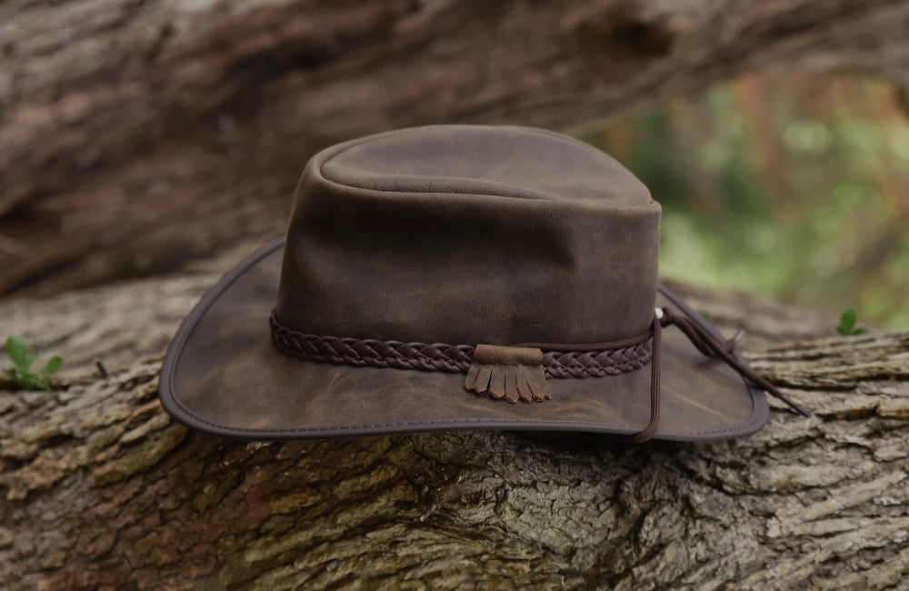 Dark brown leather outback hat with a braided band and fringed accent, resting on a rustic tree log.