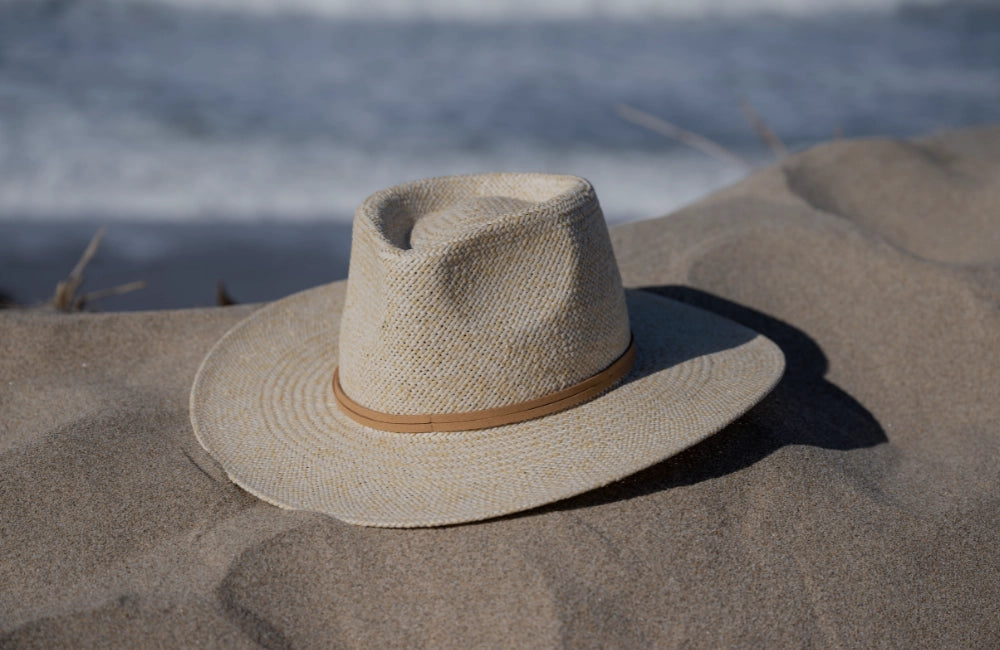 Natural woven straw wide-brim hat with a thin tan leather band, resting on a sandy beach dune near the ocean.