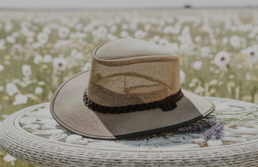 Tan breathable mesh wide-brim sun hat with a dark braided band, resting on a decorative outdoor table in a field of white flowers.