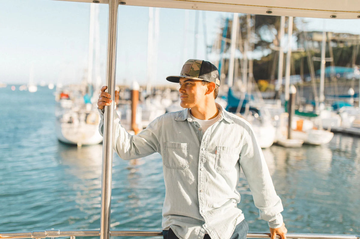 A man sitting on a ledge of a boat wearing a denim polo and a cap by American Hat Makers