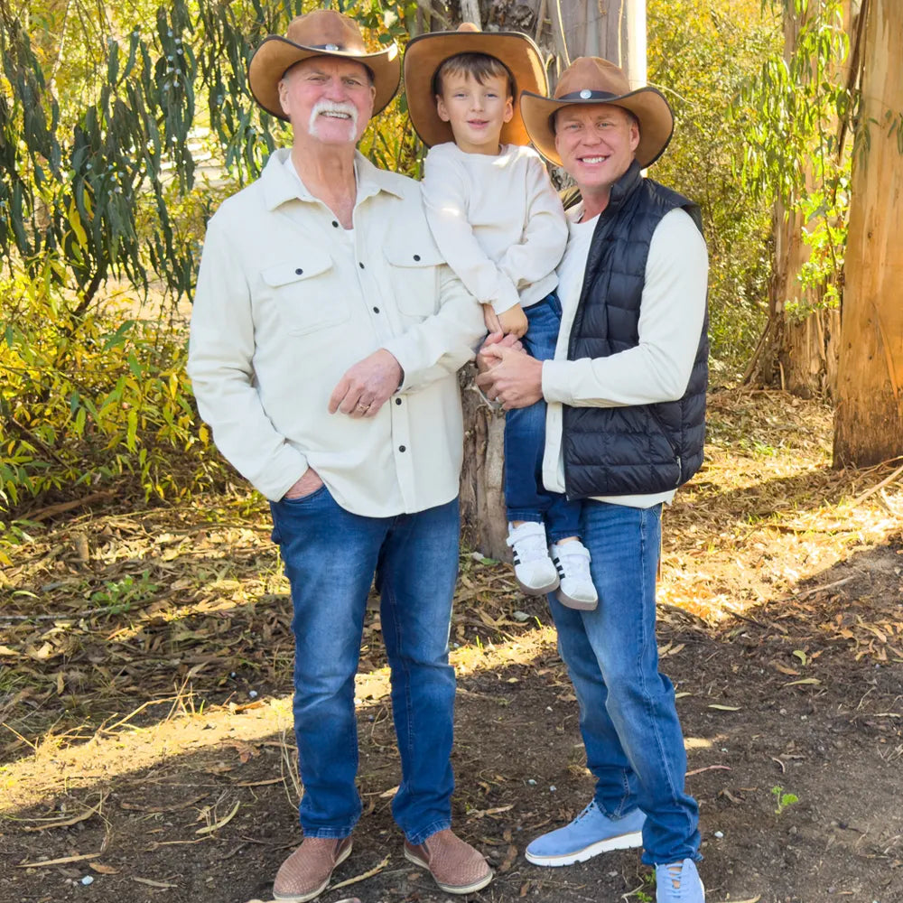 Three people wearing matching western-style outfits, including cowboy hats, cream button-up shirts, blue jeans, and boots, standing outdoors in a wooded setting with trees and leaf-covered ground.