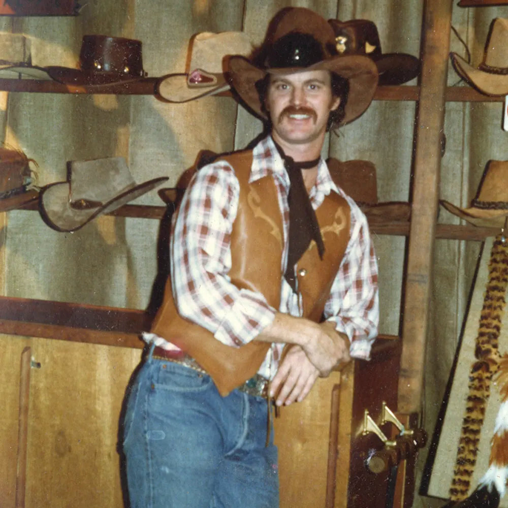 A man wearing a brown cowboy hat, brown leather vest, red and white plaid shirt, and blue jeans standing in a rustic western-themed room with shelves of cowboy hats behind him.