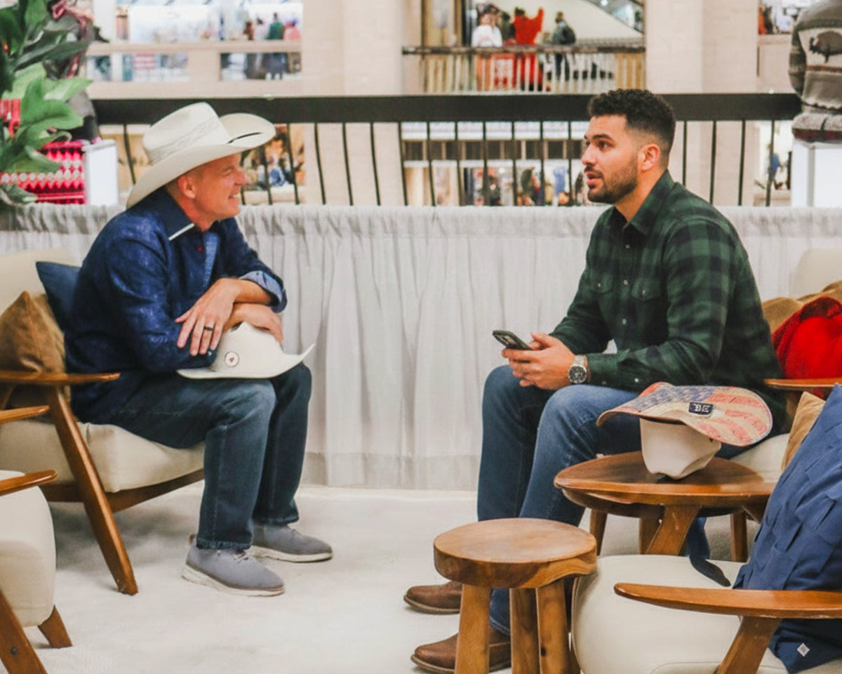 Two men sitting in a cafe or lounge setting, engaged in conversation. One man is wearing a white cowboy hat and a blue plaid shirt, while the other wears a dark green plaid shirt and holds a phone.